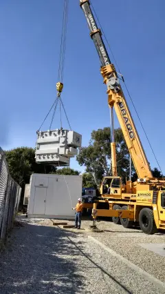 A yellow mobile crane lifting a large transformer clear of its trailer between trees and a building