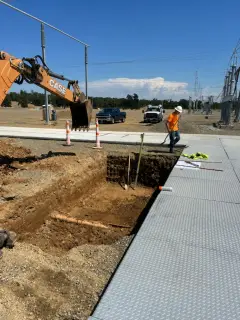 An excavator digging a narrow trench beside a concrete station road, with a worker in high-visibility clothing alongside