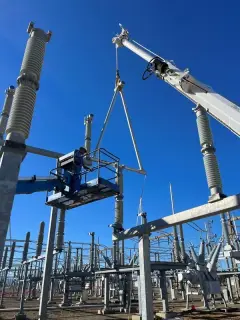 A crane hook and rigging lifting a galvanised steel structure against a clear blue sky, with substation equipment below