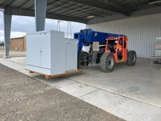 A telehandler carrying a white metal-clad switchgear cabinet on a pallet outside a steel building