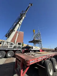 A crane lifting a steel assembly from a flatbed trailer beside a substation structure
