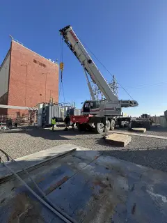 A mobile crane with its boom raised beside the tall brick wall of a pumping plant