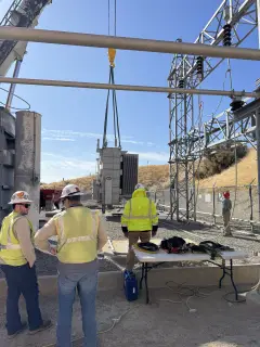 Workers in hard hats and high-visibility vests standing at a table beside the transformer pad