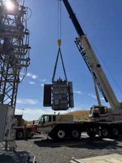 A transformer radiator assembly hanging in slings from a crane boom against a clear blue sky