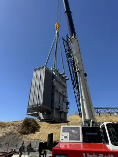 A single-phase power transformer suspended from crane rigging above a substation yard, dry hills behind