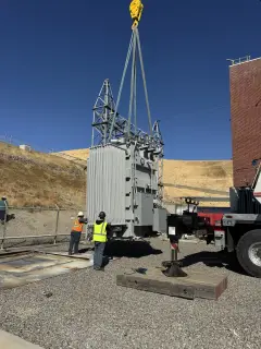 A transformer lowered towards a concrete pad while a worker in a hard hat and high-visibility vest guides it