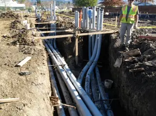 A worker standing in an open trench containing large conduits, with formwork along both edges