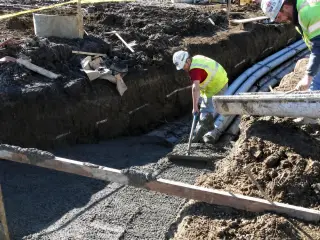 A crew placing and vibrating concrete over a run of conduits in an open trench