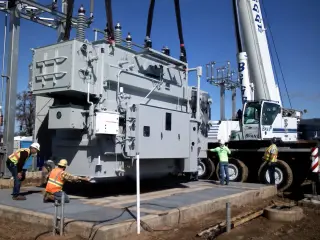 Workers guiding a large power transformer down onto a concrete pad between two cranes