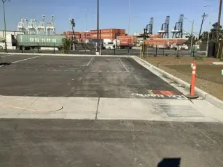A marked saw-cut line crossing a paved lot toward a fence, stacked shipping containers and ship-to-shore cranes beyond