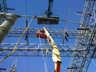 A boom-lift basket raised to a bus conductor between steel lattice structures against a clear sky