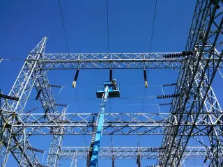 A worker in an elevated lift basket reaching up to high-voltage bus conductors and insulators against a blue sky