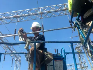 A worker high on a steel lattice structure fitting a conductor jumper, with a river valley far below