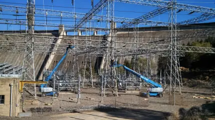 A lift working beneath the steel bus structures of a switchyard, with the face of a large concrete dam behind