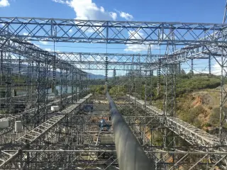 A wide view of a high-voltage switchyard with a river and wooded hills in the background