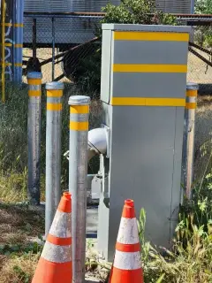 A grey electrical service pedestal with reflective banding, protected by yellow bollards and traffic cones