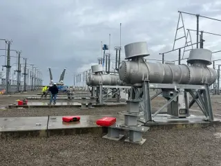 Three cylindrical capacitor units mounted on galvanised steel stands in a substation yard, with lattice towers behind
